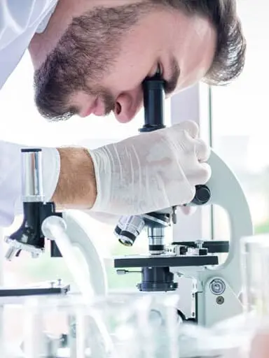 man with slicked back hair and a beard that he attempted to groom in a scientific lab with rubber gloves on looking into a microscope